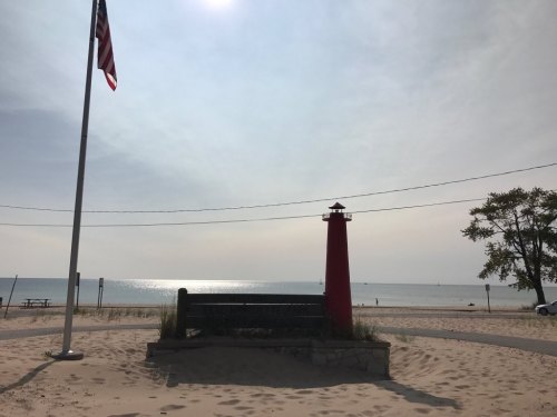 A red lighthouse stands by the beach, with an American flag waving and a calm lake in the background under a cloudy sky.