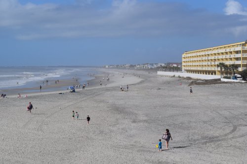 A sandy beach with people walking, a hotel in the background, and a cloudy blue sky.