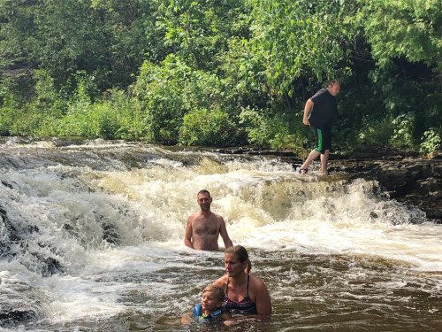 A group of people enjoying a sunny day by a waterfall, with one person wading in the water and others nearby.