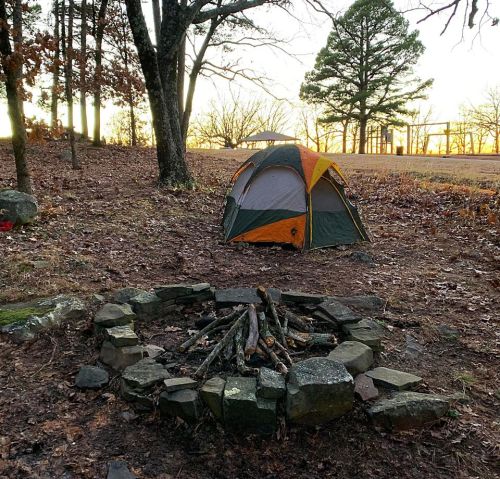 A colorful tent set up near a stone fire pit in a wooded area at sunset.