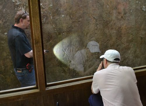 Two men examine ancient carvings on a stone wall, one using a flashlight to illuminate the markings.