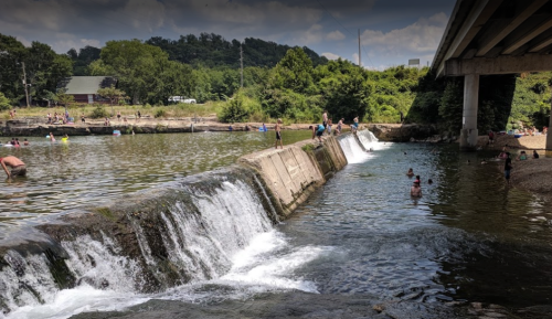 A scenic river with people swimming and relaxing, featuring a small waterfall and lush greenery along the banks.