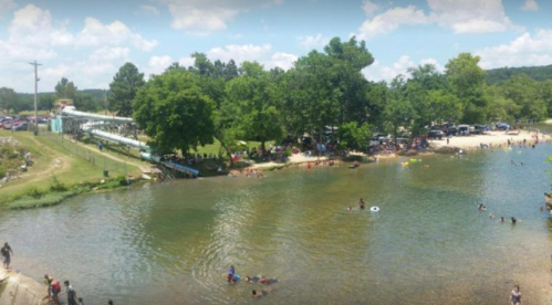 A sunny riverside scene with people swimming, lounging, and enjoying a water slide surrounded by trees.
