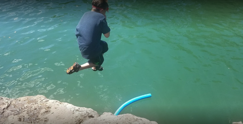 A child jumps off a rocky edge into a greenish-blue body of water, with a pool noodle floating nearby.