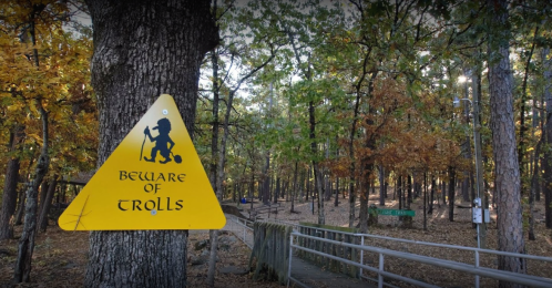 A yellow warning sign reading "Beware of Trolls" in a wooded area with autumn foliage.
