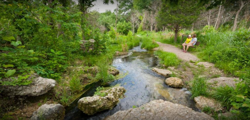 A serene stream flows through a lush green landscape, with two people sitting on a bench nearby.