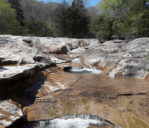A serene rocky landscape with a gentle stream flowing over smooth stones, surrounded by lush greenery.
