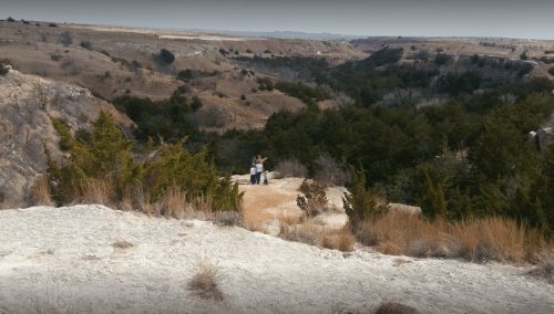 Two people stand on a rocky ledge overlooking a vast canyon filled with trees and dry terrain.