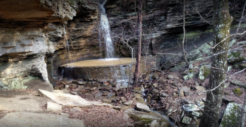 A serene waterfall cascades over rocky terrain, surrounded by trees and a stone structure in a natural setting.