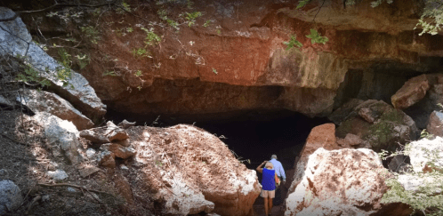 A person approaches a dark cave entrance surrounded by rocks and greenery.