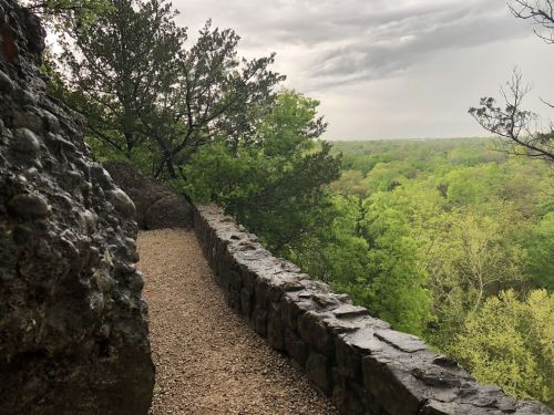 A rocky path along a stone wall overlooks a lush green landscape under a cloudy sky.