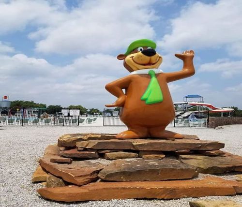 A large statue of a cartoon bear wearing a green hat and tie, standing on a stone base near a water park.