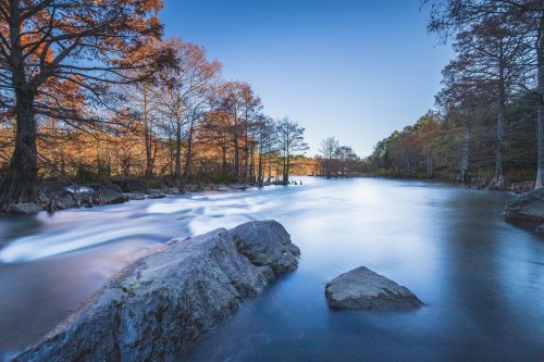 A serene river scene with smooth water, surrounded by trees with autumn foliage under a clear blue sky.