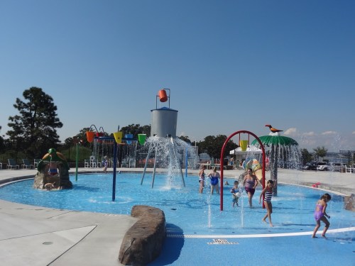 Children playing in a splash pad with water features, colorful buckets, and a sunny blue sky in the background.