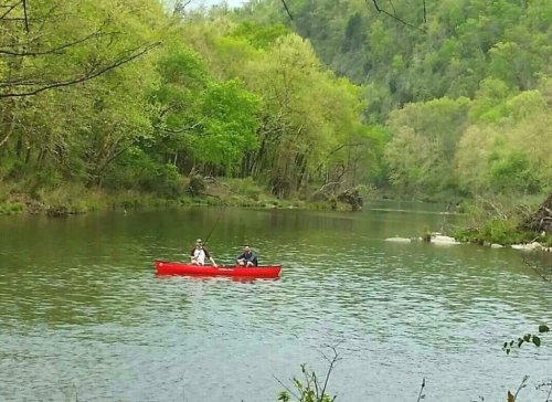 Two people fishing from a red canoe on a calm river surrounded by lush green trees.