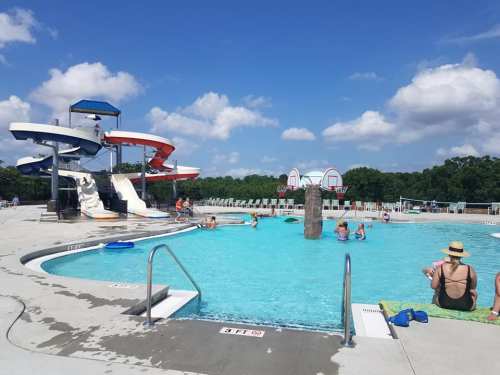 A sunny pool area with a water slide, people swimming, and basketball hoops in the background.