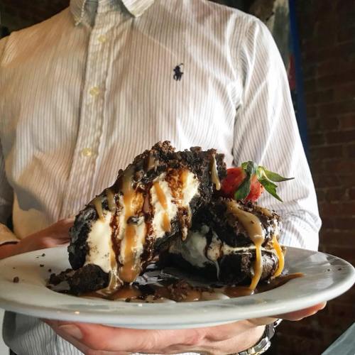A person holds a plate with a large dessert featuring ice cream, chocolate, caramel drizzle, and a strawberry on top.