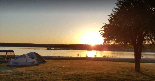 Sunset over a calm lake, with tents set up on the grassy shore and trees silhouetted against the colorful sky.