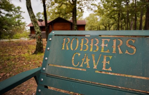 A green bench with "Robbers Cave" engraved on it, set in a wooded area with a cabin in the background.