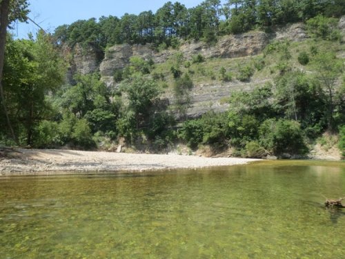 A serene river scene with clear water, sandy banks, and lush green cliffs in the background.