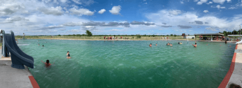 A panoramic view of a large swimming pool with people swimming and playing, surrounded by green fields and a blue sky.