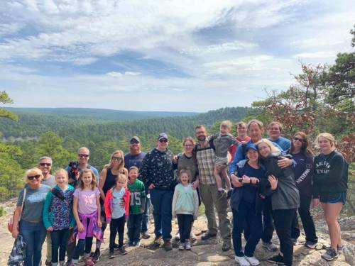 A large group of people poses together on a rocky outcrop with a scenic forested landscape in the background.