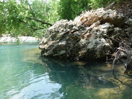 A rocky outcrop beside a clear, calm river surrounded by lush green trees.