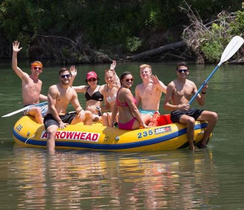 A group of seven young people wave from a yellow raft on a river, enjoying a sunny day outdoors.
