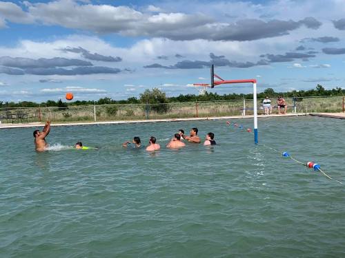 A group of people playing water polo in a pool under a cloudy sky, with one player shooting the ball towards the goal.