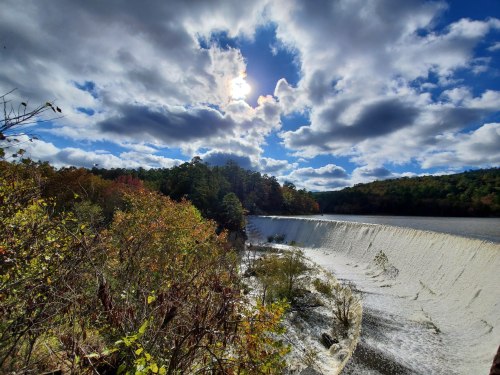 A scenic view of a waterfall under a bright sun, surrounded by trees and a cloudy blue sky.