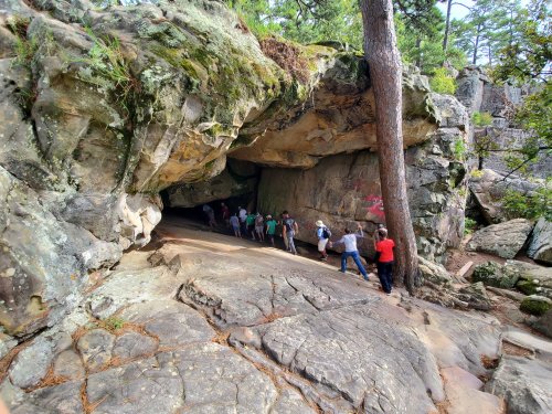 A group of people walking towards a rocky cave entrance surrounded by trees and boulders.