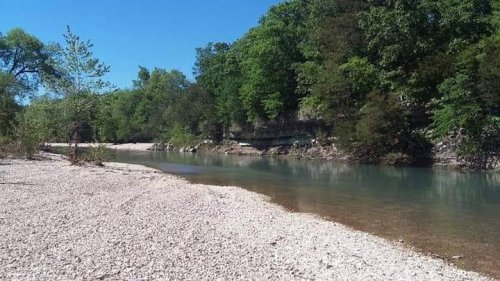 A serene riverbank scene with clear water, surrounded by lush green trees and rocky cliffs under a bright blue sky.