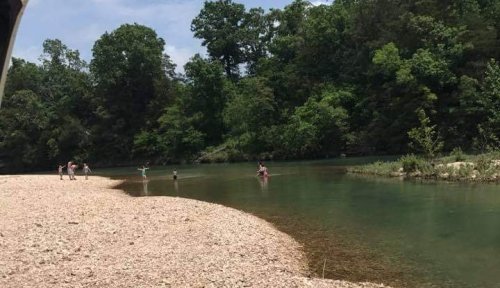 A serene river scene with people wading and playing along a pebbly shore, surrounded by lush green trees.