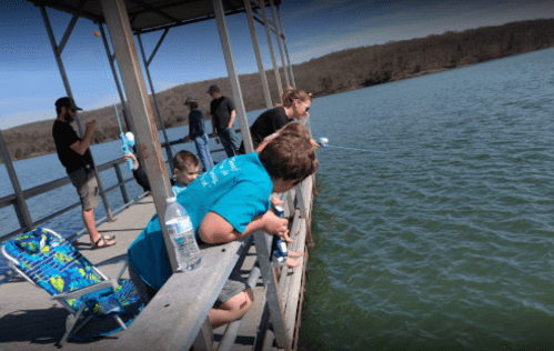 A group of people fishing from a dock by a lake on a sunny day, with children leaning over the railing.
