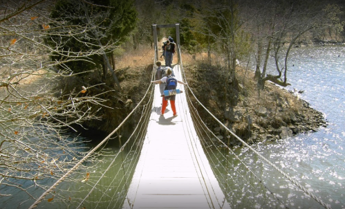 A person crosses a suspension bridge over a river, with trees and a small island in the background.