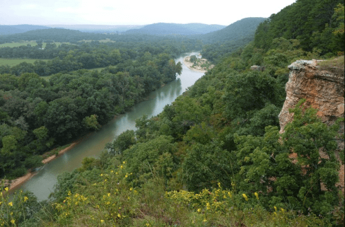 A scenic view of a winding river surrounded by lush green trees and rolling hills under a cloudy sky.