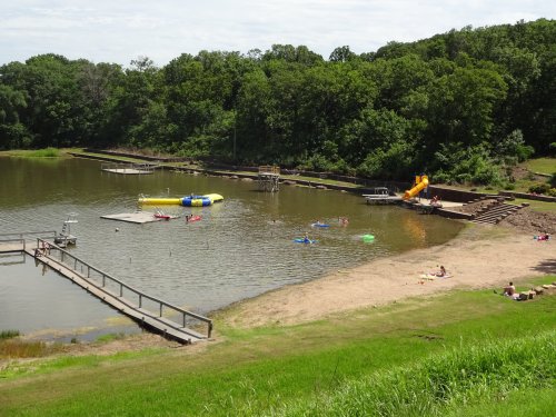 A sunny lakeside scene with people swimming, a slide, and inflatable toys, surrounded by green trees and a sandy beach.