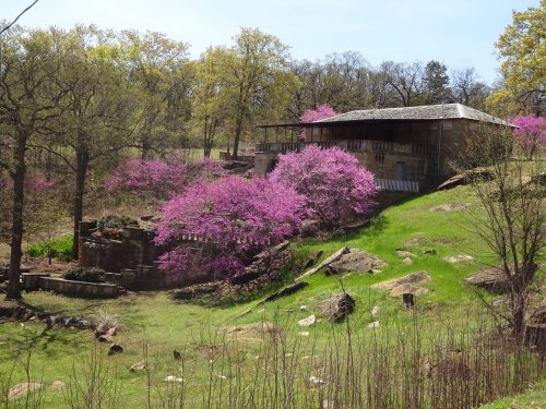 A scenic view of a hillside with vibrant pink trees and a stone house surrounded by greenery and rocks.
