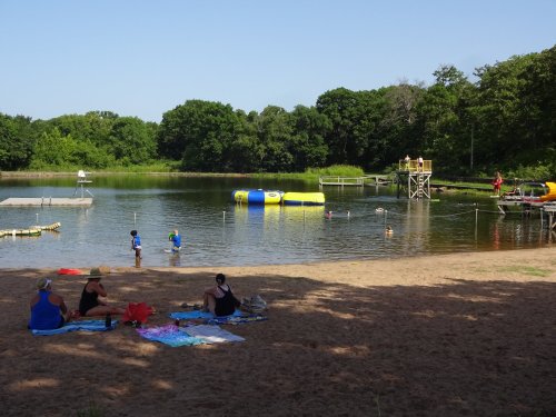 A sandy beach with people relaxing, children playing in the water, and a floating inflatable structure nearby.