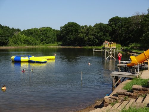 A sunny lakeside scene with a water slide, a floating trampoline, and people swimming and relaxing by the shore.