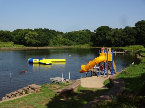 A bright yellow water slide next to a lake, with a floating inflatable and people swimming nearby. Lush greenery surrounds the area.