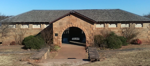 Historic stone building with a sloped roof and archway entrance, surrounded by greenery and a clear blue sky.