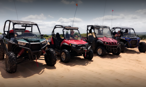 Four off-road vehicles parked on sandy terrain, with people inside and around them, under a cloudy sky.