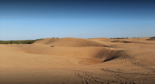 A vast desert landscape with rolling sand dunes and clear blue skies, featuring tire tracks in the sand.