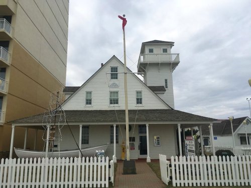 Historic building with a white facade, a tall flagpole, and a lighthouse tower, surrounded by a white picket fence.