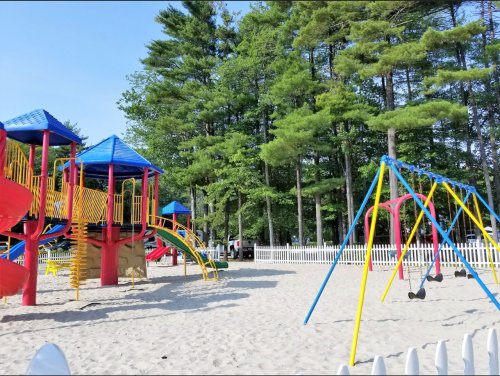 Colorful playground with slides and swings, surrounded by trees and a sandy area, enclosed by a white fence.