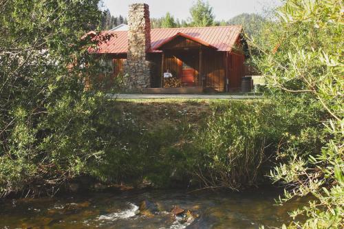 A cozy cabin with a stone chimney sits by a stream, surrounded by greenery and mountains in the background.