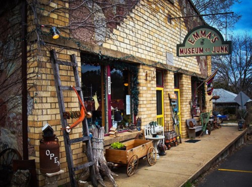 A quaint brick building with a sign reading "Emma's Museum of Junk," featuring eclectic outdoor decor and an open sign.