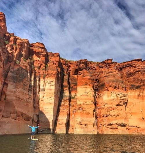 A person stands on a paddleboard in calm water, surrounded by towering red rock cliffs under a blue sky with clouds.