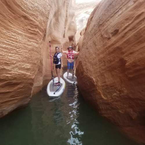 Two people paddleboarding in a narrow canyon with red rock walls and calm water.